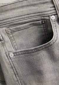 Κοντινό πλάνο γκρι τζιν από τζιν υλικό που αναδεικνύει την εμπρόσθια τσέπη με μεταλλικά καρφιά και εμφανή διπλά ραφές.