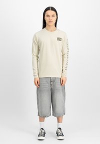 Camisa de manga larga color beige claro con un parche en el pecho, combinada con pantalones cortos de mezclilla desgastados hasta la rodilla y zapatillas negras.