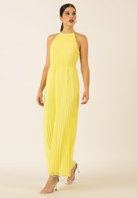 Robe dos-nu plissée en jaune vif. Design fluide avec une taille élastique. Associée à des talons argentés à brides. Texture lisse.