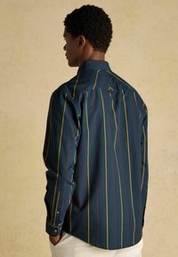 Muž otočený chrbtom, oblečený v tmavomodrej košeli s vertikálnymi žltými a červenými pruhmi a bielych nohaviciach, pred jednoduchým béžovým pozadím.