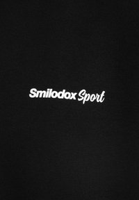 Tessuto nero con una consistenza morbida, caratterizzato da un ricamo bianco con il testo "Smilodox Sport" in un font moderno. Design semplice e pulito.