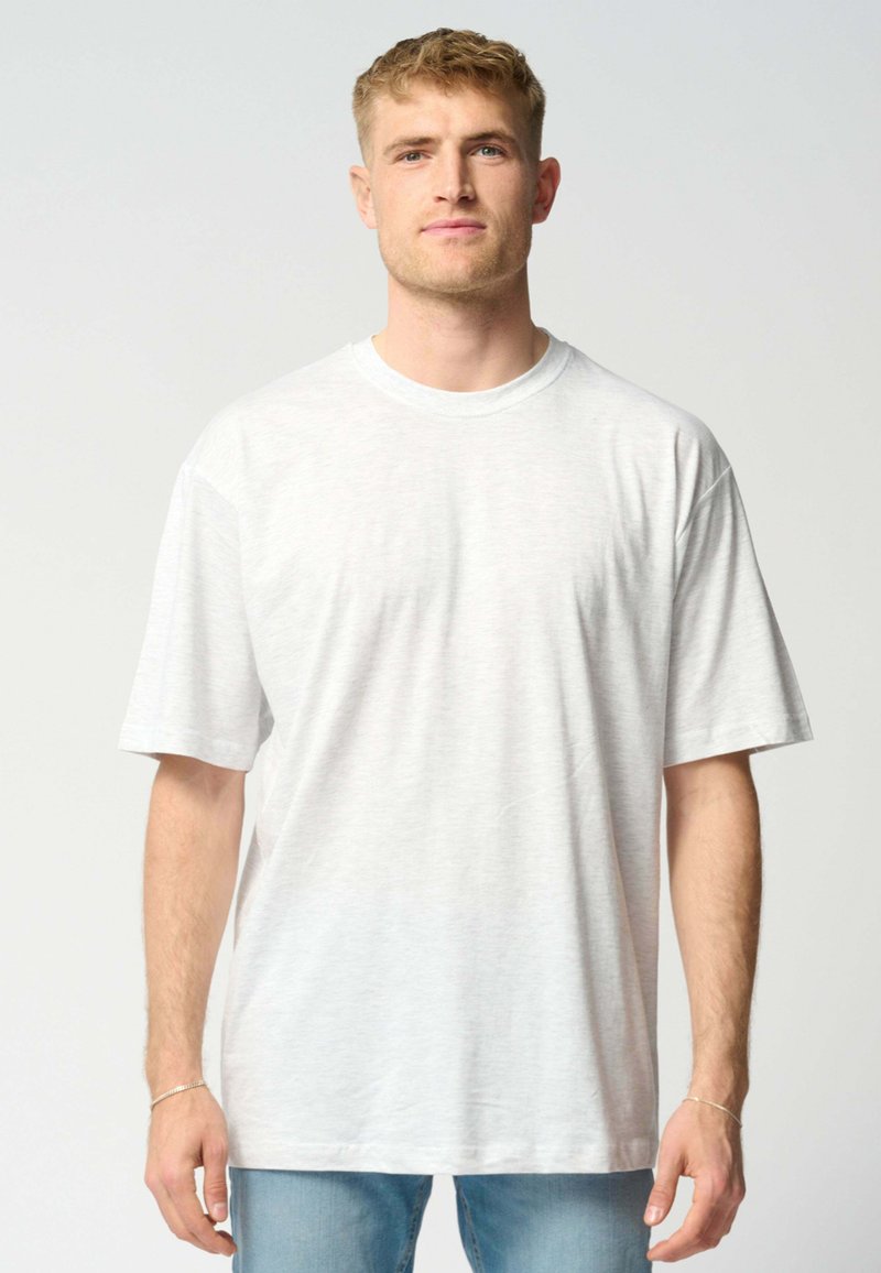 En blond mand iført en simpel hvid oversized T-shirt og lyseblå jeans, der står og vender fremad mod en lys grå baggrund.