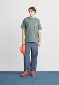 Πράσινο βαμβακερό μπλουζάκι με τσέπη στο στήθος, συνδυασμένο με μπλε τζιν παντελόνι, πορτοκαλί καπέλο και καφέ δετά παπούτσια, που στέκεται σε μπλε πάτωμα.