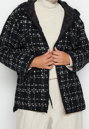 Person trägt eine schwarz-weiß karierte Jacke mit zwei Fronttaschen und richtet den Reißverschluss über einem cremefarbenen Rollkragenpullover und hellen Hosen.