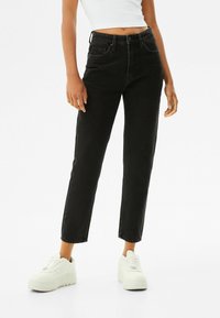 Jeans negros de cintura alta con corte recto, cinco bolsillos y una textura ligeramente deslavada, combinados con zapatillas blancas de caña baja.