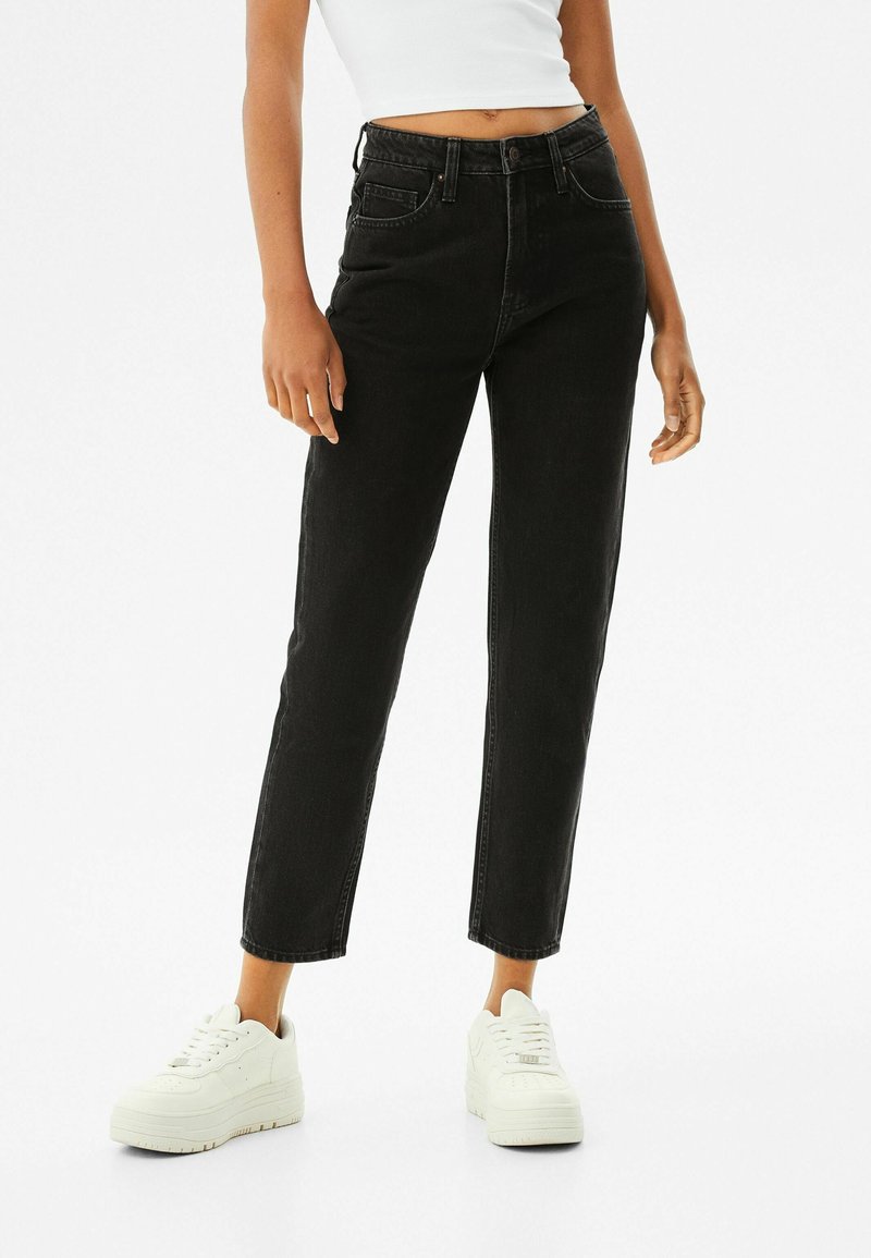 Jeans negros de cintura alta con corte recto, cinco bolsillos y una textura ligeramente deslavada, combinados con zapatillas blancas de caña baja.
