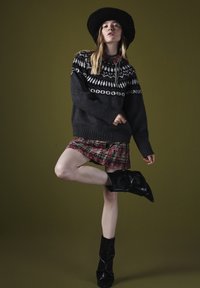 Grauer gestrickter Pullover mit weißen gemusterten Akzenten, rotem Karorock und schwarzen kniehohen, gehäkelten Stiefeln, vor einem grünen Hintergrund.