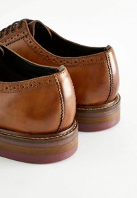 Pantofi de dress din piele maro cu un finisaj neted, având detalii brogue, vârful rotund și un toc stratificat cu dungi contrastante.