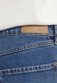 Blue denim jeans featuring a tan leather patch with "ESPRIT" embossed. Notable details include yellow stitching and a smooth texture.