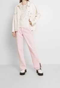 Cremefarbene Jeansjacke über einem weißen Pullover mit Verzierungen, kombiniert mit hellrosa, weitem Hosen und schwarzen Plateau-Schuhen.