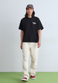 T-shirt en coton noir avec logo sur la poitrine gauche, associé à un pantalon crème à rayures et des baskets rouges. L'arrière-plan est pâle et minimal.