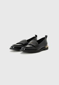 Zwarte leren instappers met een glanzende afwerking, decoratieve stiksels en een goudkleurig accent op de hak. Lage, platte zool.