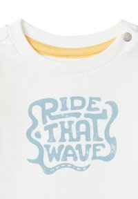 Witte katoenen shirt met een ronde halslijn en een gele rand. Centraal staat in blauwe tekst "RIDE THAT WAVE" in een speelse lettertype.