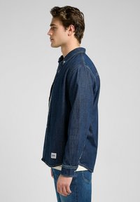 Chaqueta de mezclilla en azul oscuro, con cierre de botones en la parte delantera, cuello clásico, mangas largas y una etiqueta de marca en el dobladillo inferior.