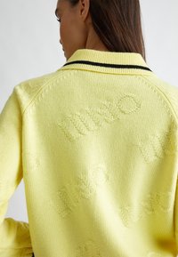 Maglione giallo a maglia con un motivo testurizzato in rilievo con la parola "WHO" e un colletto a righe nere.