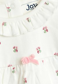 Biela detská šaty s malými ružovými a zelenými kvetinovými vzormi, volánovým golierom a mäkkou ružovou zamatovou mašľou na hrudi.