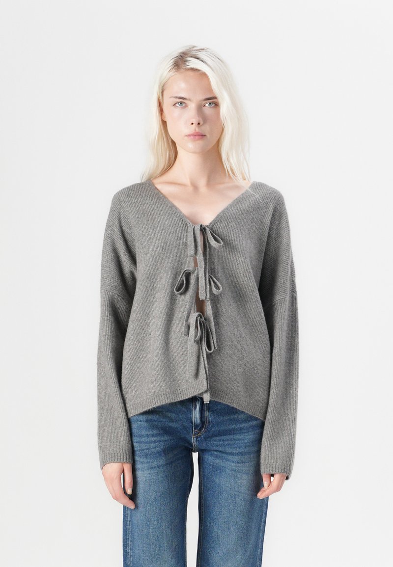 Γκρί πλεκτό πουλόβερ με βαθύ V-λαιμό, κορδόνια μπροστά και φαρδιά μανίκια. Συνδυασμένο με μπλε τζιν. Υφασμάτινη υφή, χαλαρή εφαρμογή.