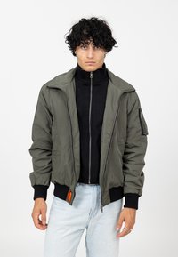 Veste bomber vert olive avec fermeture éclair à l'avant, poignets côtelés et étiquette orange. Portée sur un haut noir à col montant, avec une coupe classique.