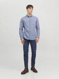 Camisa azul de botones con un bolsillo en el pecho, combinada con vaqueros oscuros y botas de tobillo marrones. La camisa presenta un cuello y un diseño de corte clásico.