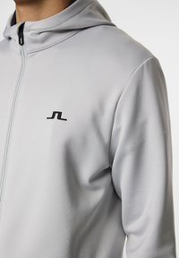 Hellgraue Zip-Up-Hoodie aus glattem Material, mit hohem Kragen, Frontreißverschluss und kleinem schwarzem Logo auf der Brust.