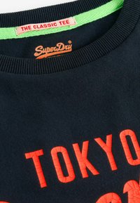 Tumesinine puuvillane t-särk ümarate kraega, millel on pragunenud punane "TOKYO" tekst ja SuperDry logo laimirohelise kaelana.