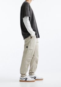 Gray short-sleeve shirt over a white long-sleeve shirt, paired with beige cargo pants and white sneakers with black accents.