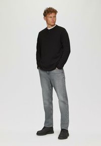 Schwarzes, langärmliges Henley-Shirt mit zwei Knöpfen auf der Vorderseite, kombiniert mit hellgrauen, geraden Jeans und schwarzen Schuhen, vor einem neutralen Hintergrund.