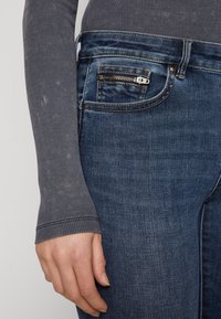 Jeans de mezclilla azul oscuro con un bolsillo lateral con cierre y costuras en contraste, combinados con una camiseta de manga larga ajustada de canalé gris.