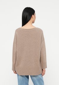 Oversized beżowy sweter, miękka tekstura, szeroki dekolt, ściągacze przy mankietach i na dole, posiada konstrukcję typu high-low. Noszony z jasnoniebieskimi dżinsami.