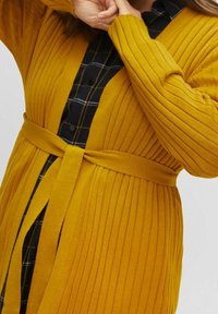 Musztardowożółty prążkowany kardigan z paskiem do wiązania, noszony na czarnej koszuli w kratę. Posiada długie rękawy i teksturowaną tkaninę.