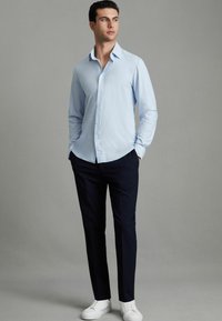 Hellblaues Hemd mit spitzem Kragen, kombiniert mit dunkelblauen Hosen und weißen Sneakers. Glattes Material, taillierte Passform.