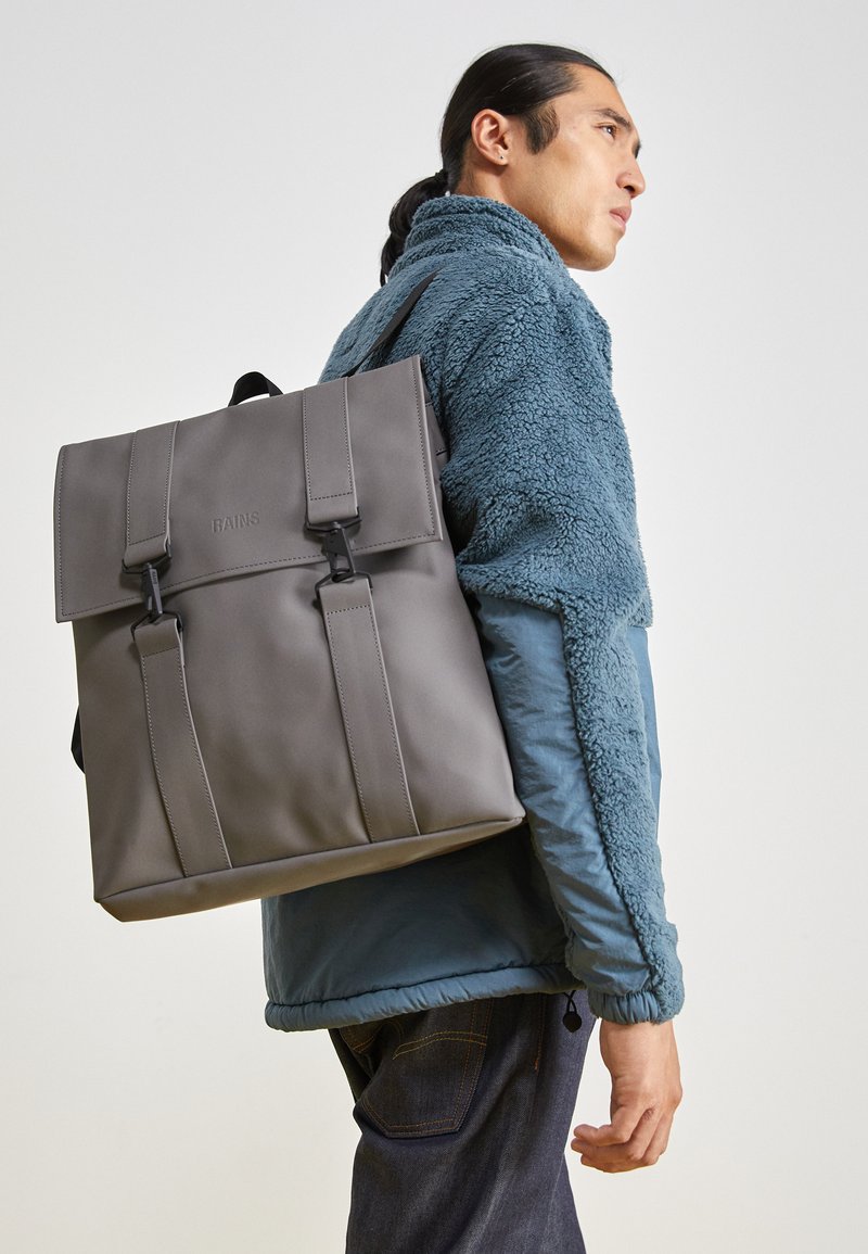 Mochila impermeable gris con un diseño estructurado, que presenta correas frontales dobles y un cierre con solapa, llevada sobre una chaqueta de felpa verde azulado.