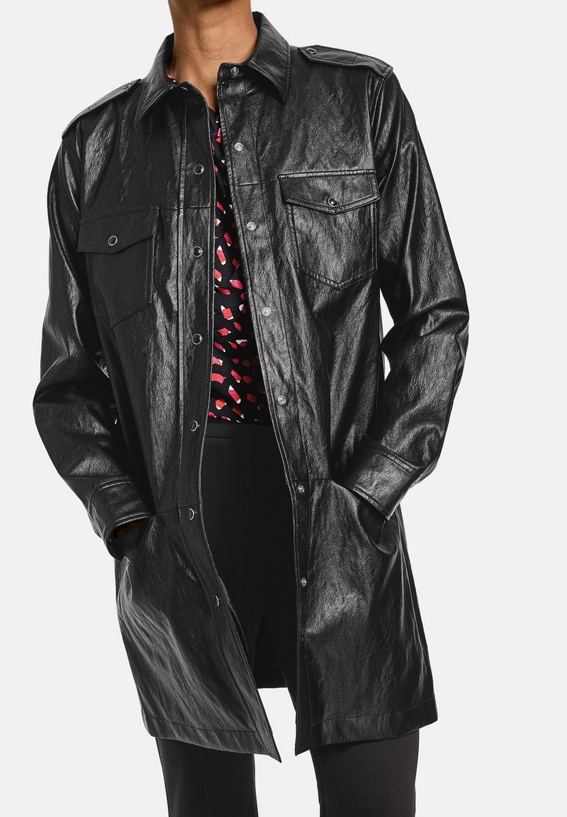 Personne portant un long manteau en cuir noir avec des boutons-pression par-dessus un haut noir imprimé de lèvres rouges et blanches, les mains dans les poches du manteau.