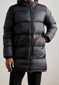 Homme portant un manteau matelassé noir avec capuche, fermeture éclair à l'avant et poches latérales zippées, une main dans la poche et l'autre détendue.