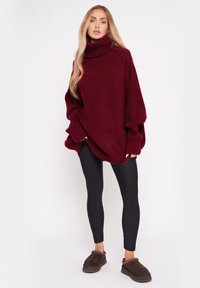 Burgundowa, oversize sweter z wysokim golfem, połączony z czarnymi legginsami i brązowymi butami wsuwanymi z dekoracyjnym przeszyciem.