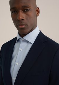 Mornarsko suknjo z izrezanim ovratnikom in svetlo modro teksturirano srajco pod njim, ki ima bele gumbe in majhen detajl žepa na prsih.