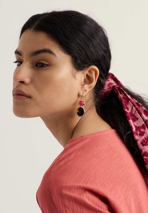 Goudkleurige oorbellen met een rode steen op een basis in schildpadvorm, gedragen met een roze top en een haarband met bloemenpatroon.