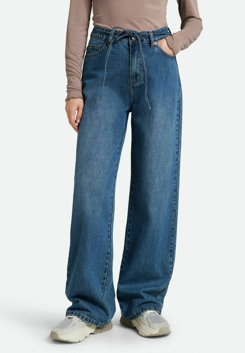 Jeans de denim de pierna ancha en azul medio con cintura alta y cierre con cordón. Cuenta con bolsillos frontales y un ajuste suelto y relajado.