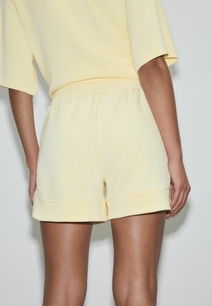 Persoană purtând pantaloni scurți galbeni deschis cu talie elastică și top larg cu mâneci scurte asortat, văzută din spate pe un fundal simplu.