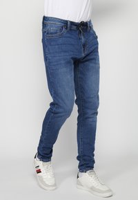 Joggers de mezclilla en azul oscuro con corte ajustado, cintura con cordón y bolsillos delanteros. Combinados con zapatillas blancas que cuentan con detalles en negro y rojo.