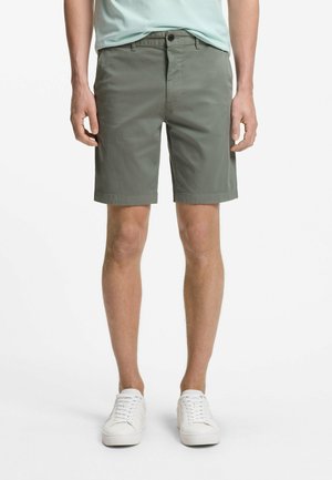 Mees, kes kannab oliivrohelisi põlveni ulatuvaid lühikesi pükse, helerohelist t-särki ja valgeid tossusid, seisab valge tausta ees.