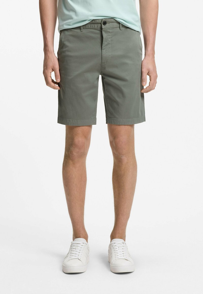 Άνδρας που φοράει χακί σορτς μέχρι το γόνατο, μπλουζάκι σε μέντα πράσινο χρώμα και λευκά αθλητικά παπούτσια, στέκεται μπροστά σε λευκό φόντο.