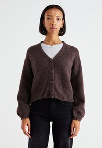 Hnedý pletený cardigan s výstrihom do V, piatimi tmavými gombíkmi, rebrovaným pásom a dlhými rukávmi, nosený cez svetlošedú košeľu.