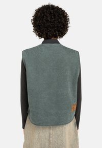 Groene canvas bodywarmer met een afgeronde zoom en een patchdetail. Heeft een zwarte kraag en wordt gedragen over een zwart shirt met lange mouwen.