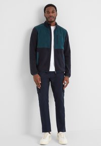 Fleece jacket in dark navy and teal, with a full zip, stand collar, front pockets, and a zippered chest pocket. Worn with dark pants and sneakers.