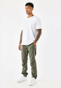 Camiseta blanca de manga corta, pantalones joggers verde oliva con puños elásticos y zapatillas blancas. Presenta tatuajes en los brazos y un diseño minimalista.