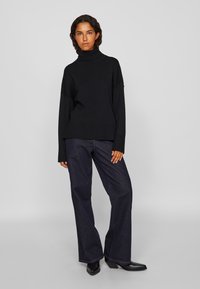 Μαύρο πουλόβερ με λαιμόκοψη τύπου τσόκερ και χαλαρή εφαρμογή, συνδυασμένο με σκούρο μπλε φαρδύ παντελόνι τζιν και μαύρες μυτερές μποτες μέχρι τον αστράγαλο. Λείο υλικό και μινιμαλιστικός σχεδιασμός.