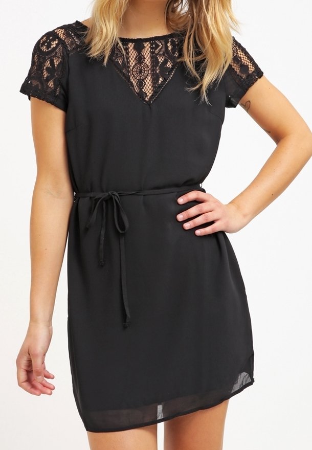 Femme portant une robe noire courte avec manches en dentelle et décolleté, nouée à la taille, debout avec une main sur la hanche.