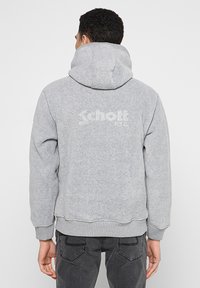 Grijze capuchontrui met zachte textuur, geribbelde manchetten en zoom. Heeft een vaag logo "Schott NYC" op de achterkant. Klassieke, casual pasvorm.