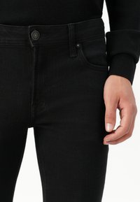 Primer plano de unos jeans de mezclilla negros combinados con una blusa negra de manga larga, mostrando la cintura, el botón y una mano relajada al costado.